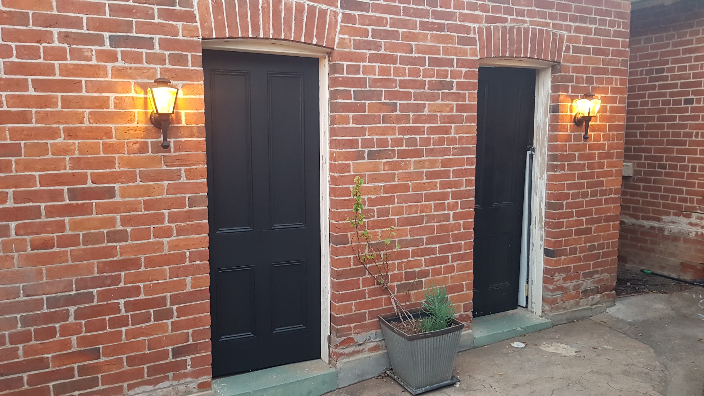 Two coach lanterns lit either side of black doors on a red brick wall
